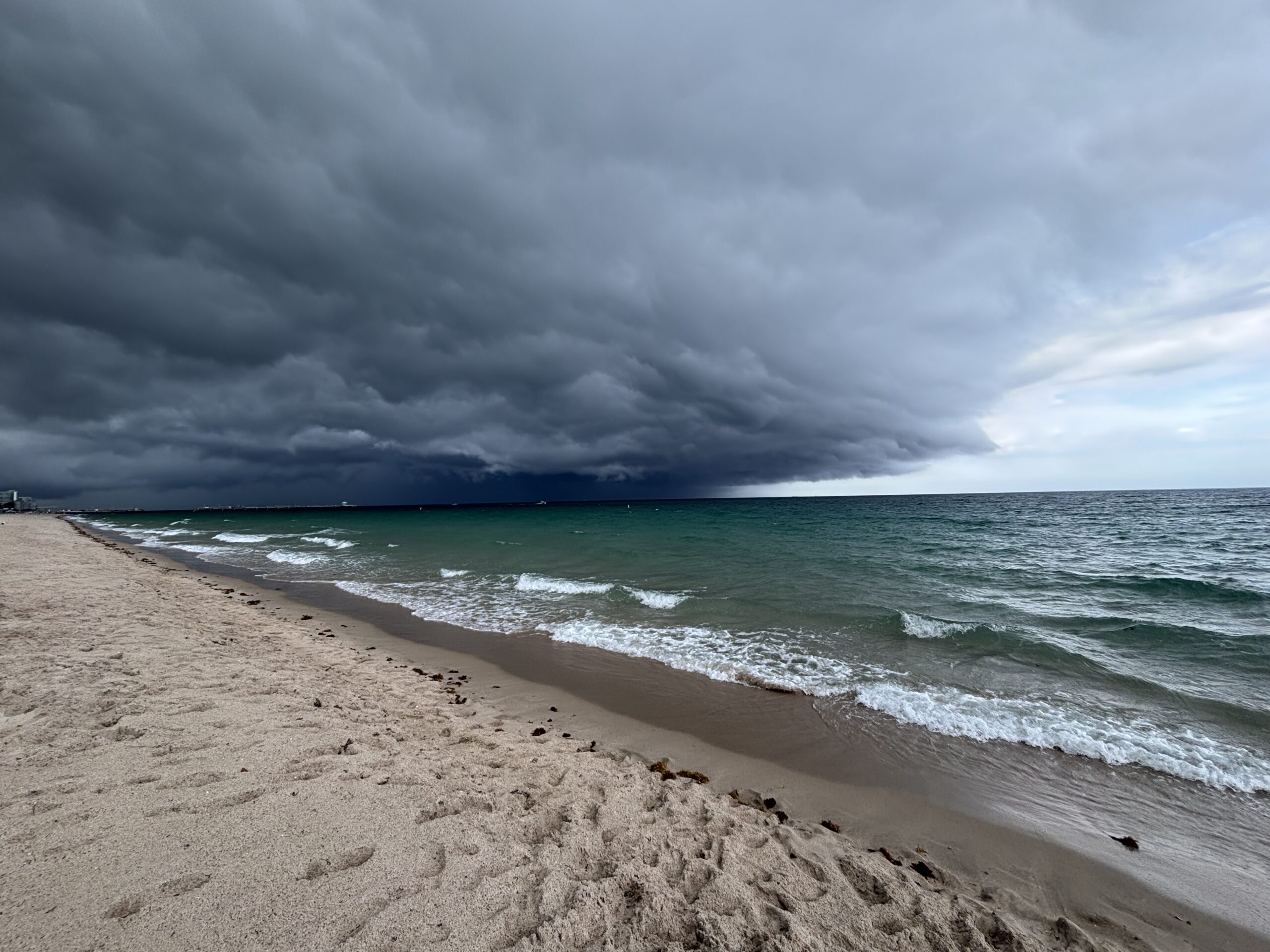 Dark storm clouds rolling over the ocean along a sandy beach, with waves approaching the shore and a dramatic sky forming in the distance.